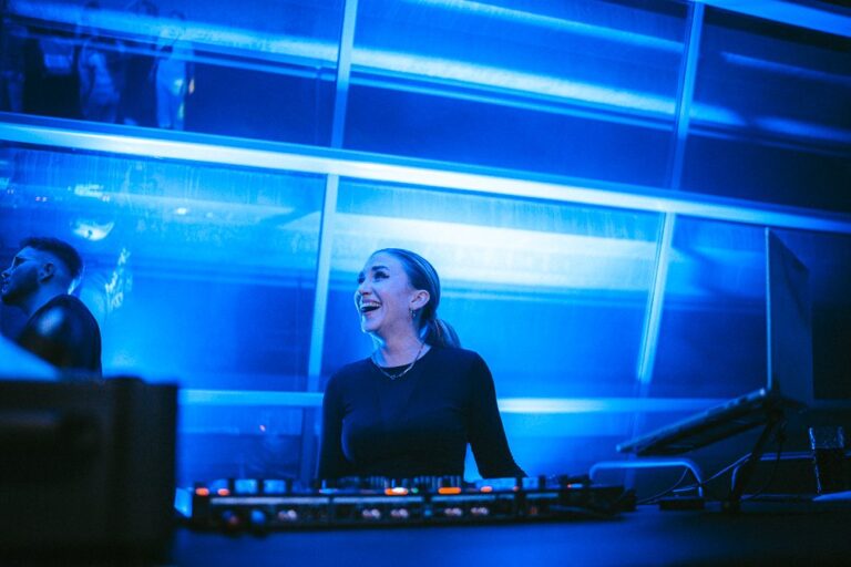 DJane Polly lacht am DJ-Pult bei einem Abendevent in blauem Licht
